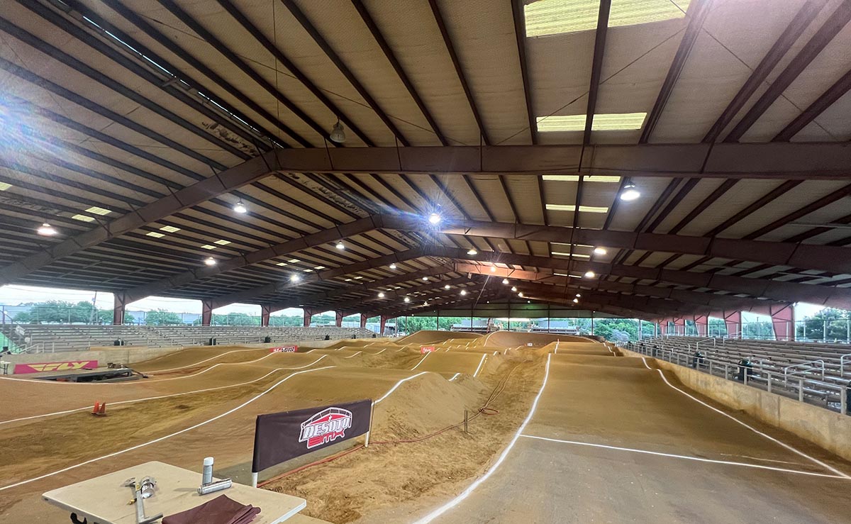 A wide view of DeSoto BMX Track in Texas, featuring a large, covered facility with a well-lit, hard-packed dirt track, massive berms, and high jumps under a clear blue sky.