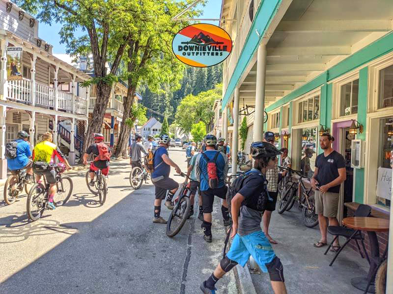 A street view of a road lined with stores, with MTB riders gathered outside, shopping and resting with their bikes.