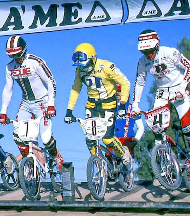 BMX riders from the 70s-80s racing in colorful jerseys, old-school BMX pants, and helmets.